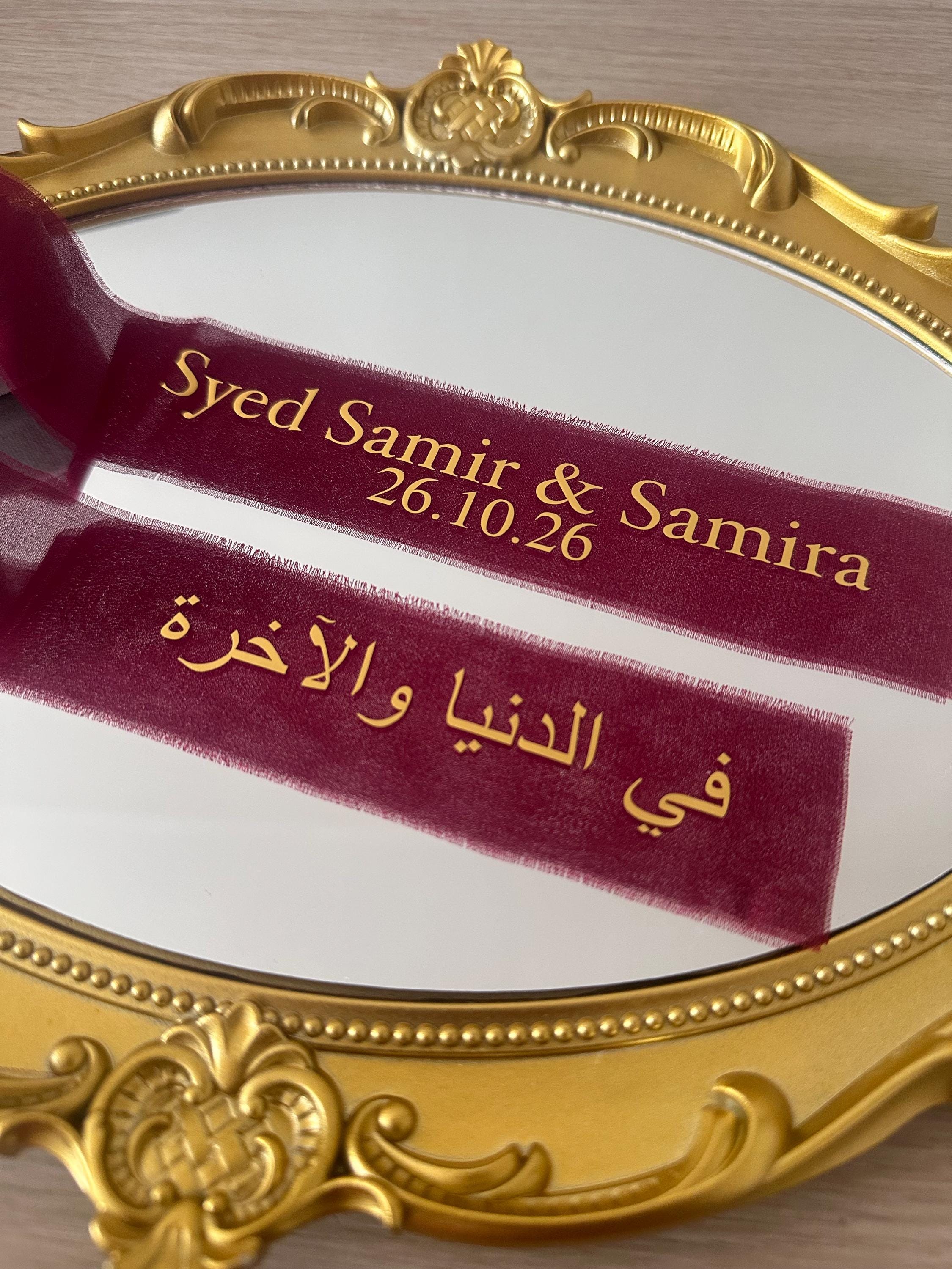 Burgundy personalised ribbons with gold names, a date and Arabic wording on a gold ring plate.